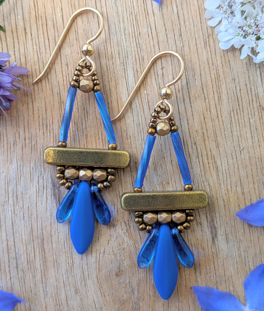 Blue and gold earrings with long blue dagger beads at the bottom, on a wood background, with flowers.