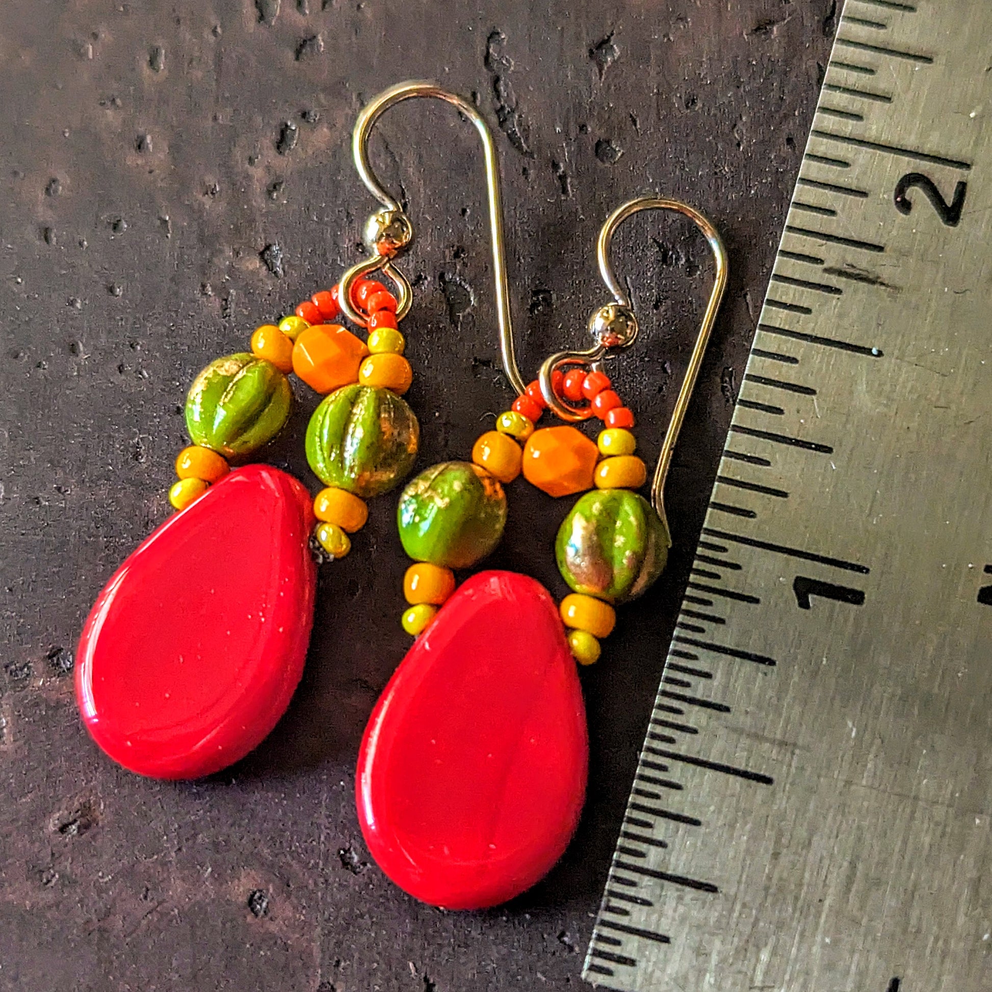 Colorful beaded earrings with red teardrop-shaped beads accented with green, yellow and orange beads on a textured surface with a ruler for scale.