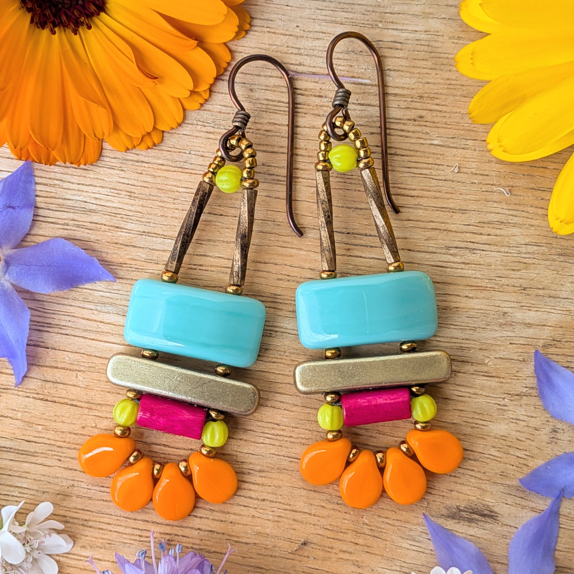Colorful earrings with turquoise rectangles on top, a gold bar in the middle and a magenta tube with a swag of petal-like orange beads on a wooden surface with flowers.