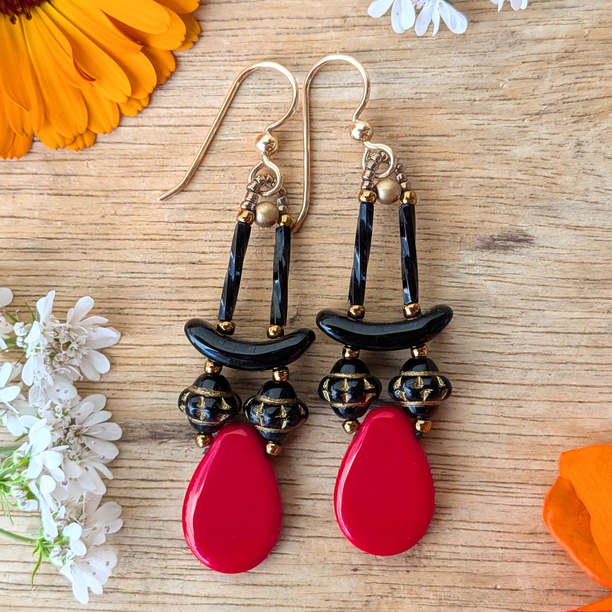 Red and black earrings on a wood background with flowers. The earrings are composed of large red teardrop sat the bottom with a pair of gold-patterned, textured black beads and black arcs on top. 