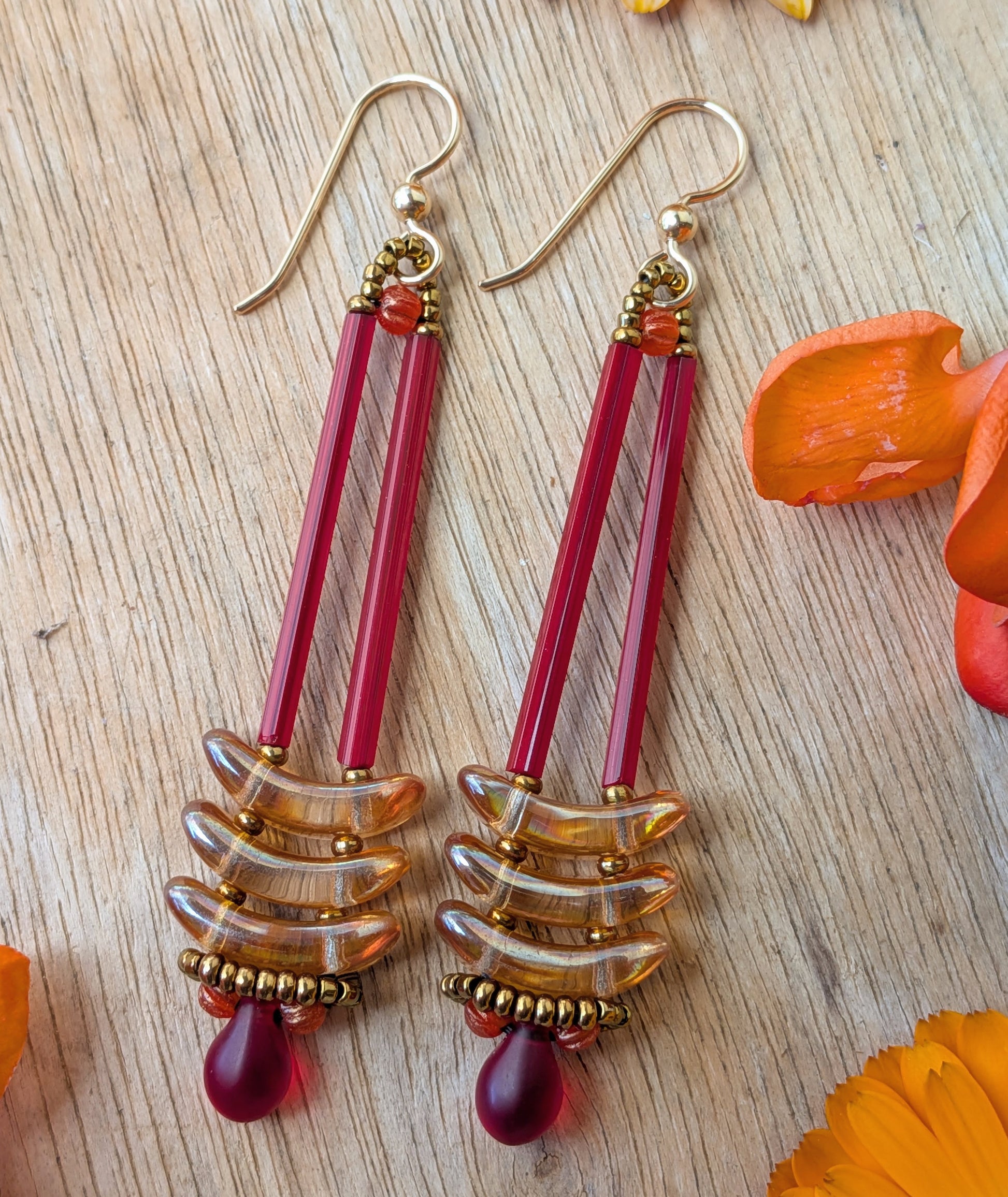 Long red an apricot earrings on a wood background with orange flowers.