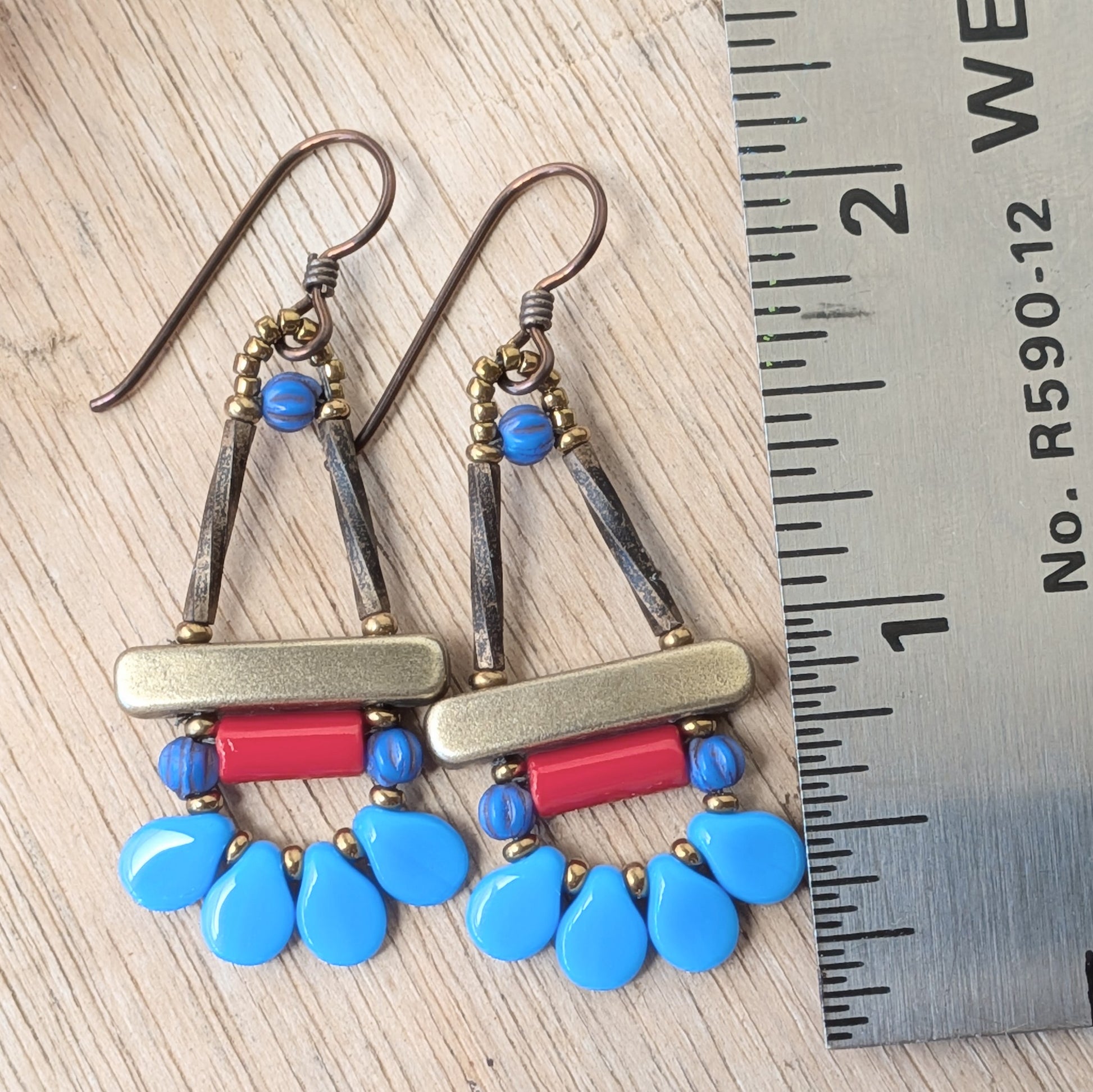 Blue and red earrings with petal shaped beads around the bottom, on a wood background with flowers and a ruler for scale. 