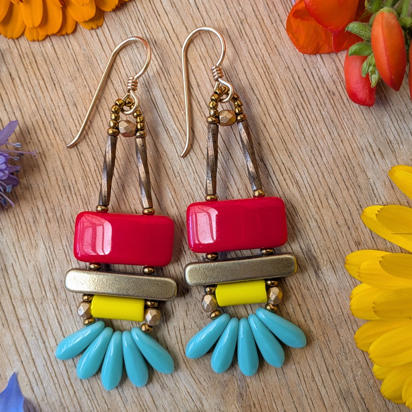 colorful earrings on a wood background with flowers. The earrings each have a red rectangle stacked on top of a brass-colored bar. Under the bar there is light yellow tube, five small turquoise daggers and tiny gold accents.