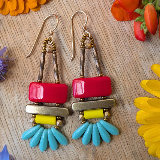 colorful earrings on a wood background with flowers. The earrings each have a red rectangle stacked on top of a brass-colored bar. Under the bar there is light yellow tube, five small turquoise daggers and tiny gold accents.