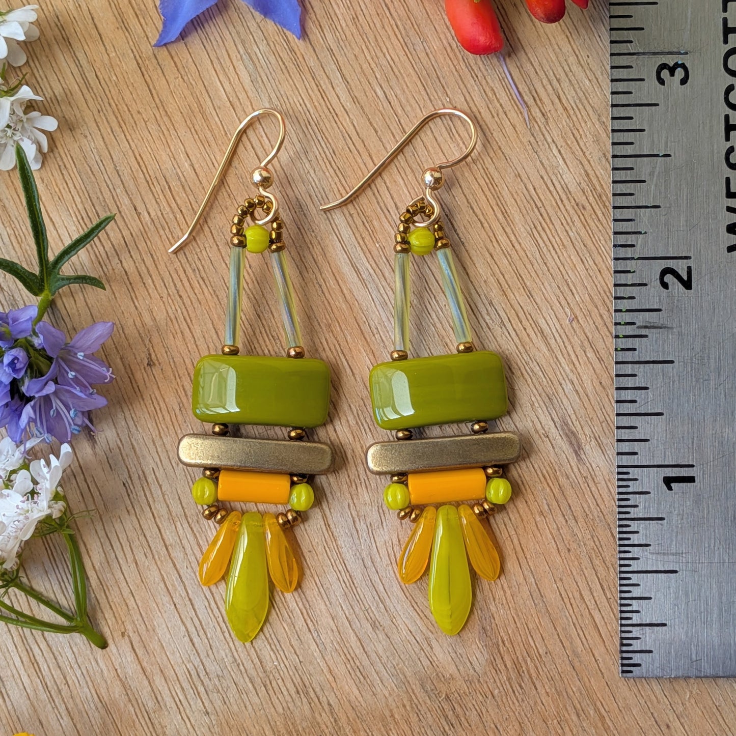 Colorful olive green, orange and chartreuse earrings on wood background with a ruler for scale. There is an avocado green rectangle on top, stacked above a gold bar. Below the bar is an orange tube and a swag of orange and chartreuse dagger beads.
