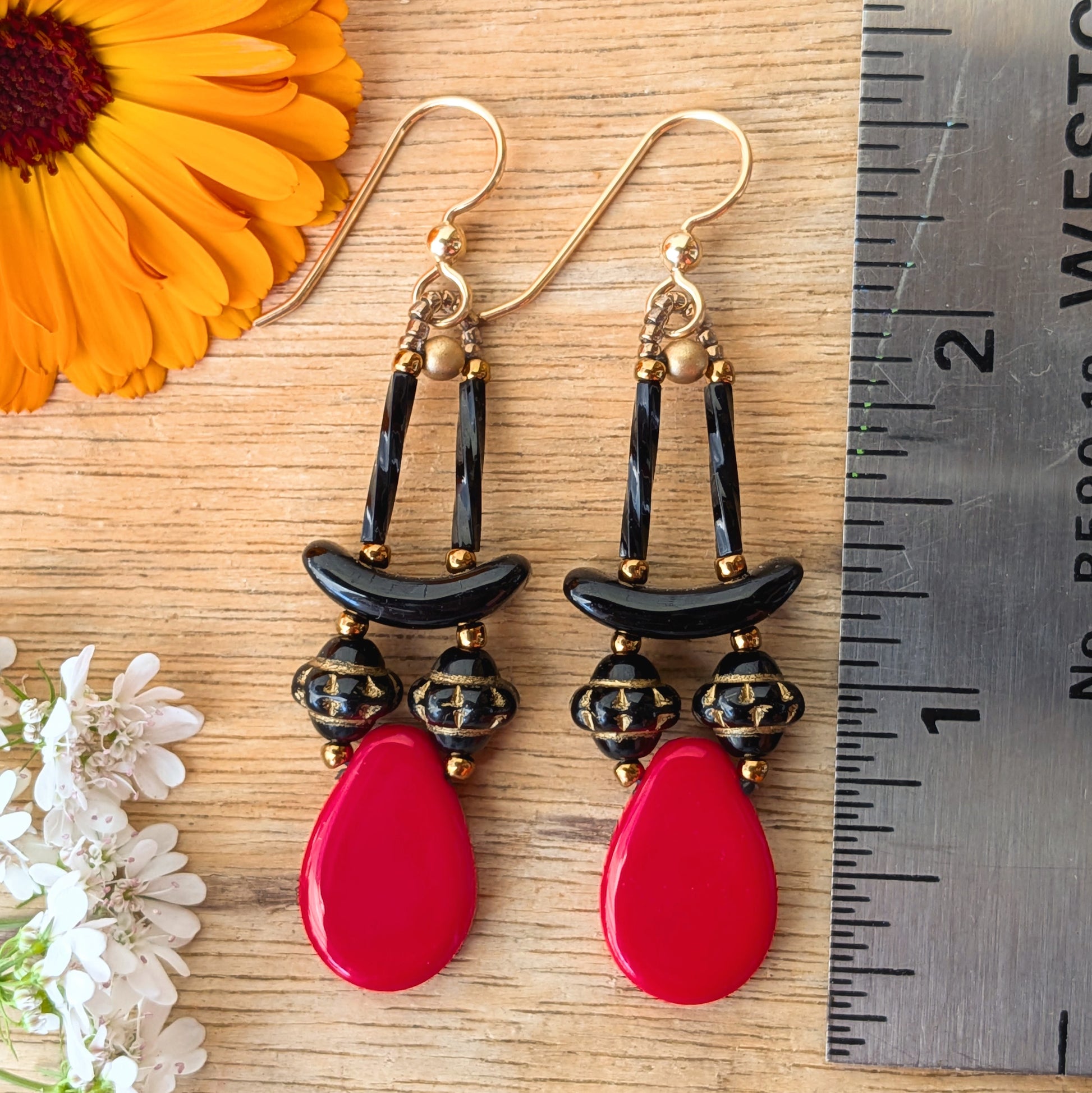 Red and black earrings on a wood background with flowers and a ruler for scale. The earrings are composed of large red teardrop sat the bottom with a pair of gold-patterned, textured black beads and black arcs on top. 
