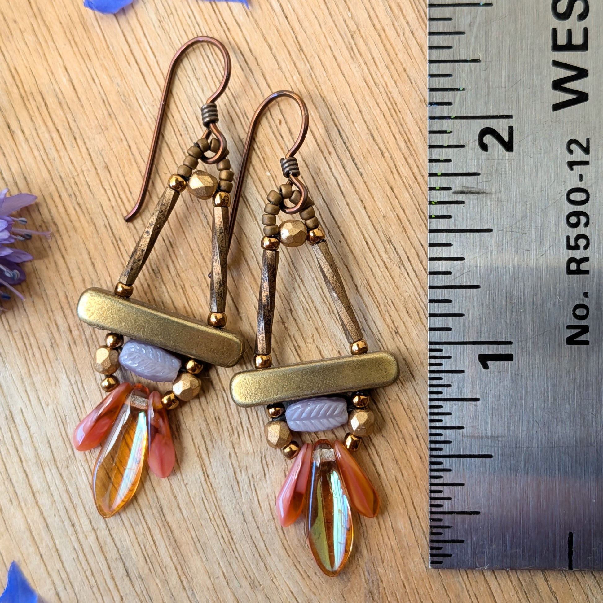 A pair of gold, lilac and peachy-pink earrings on a wood background with flowers and a ruler for scale.