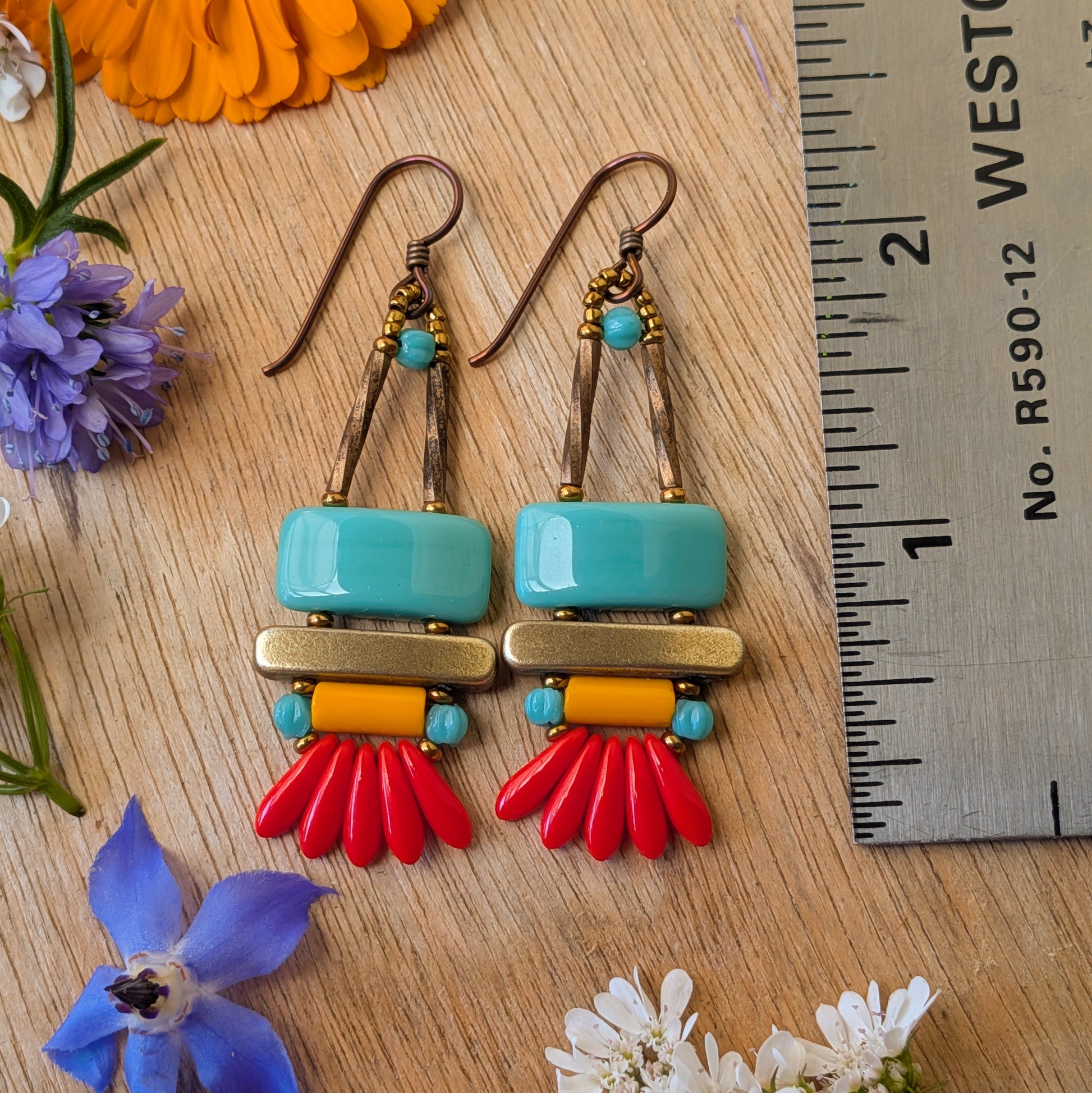 colorful earrings on a wood background with a ruler for scale. The earrings each have a wide turquoise rectangle stacked on top of a brass colored bar. Under the bar there is light orange tube, five small red daggers and tiny turquoise accents.