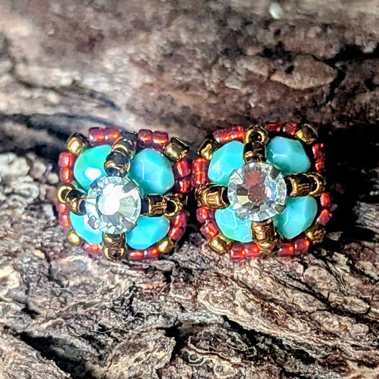 Four-lobed stud earrings made with larger turquoise and smaller red beads and a rhinestone center, on a wood bark background.