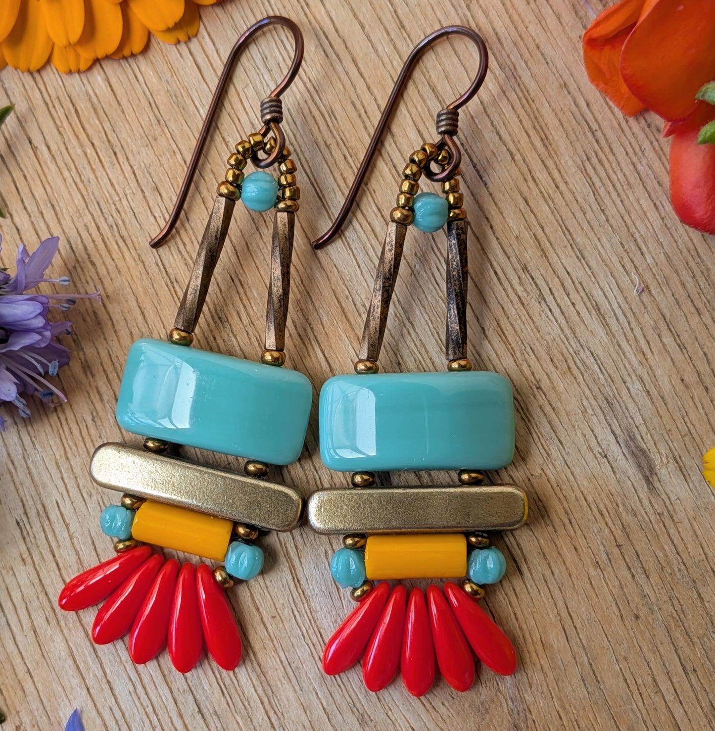 colorful earrings on a wood background. The earrings each have a wide turquoise rectangle stacked on top of a brass colored bar. Under the bar there is light orange tube, five small red daggers and tiny turquoise accents.