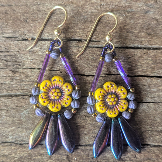 A pair of earrings with yellow flowers and a fringe of iridescent purple beads on a wooden surface
