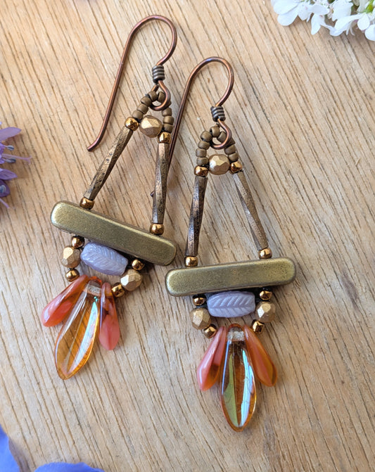 A pair of gold, lilac and peachy-pink earrings on a wood background with flowers.