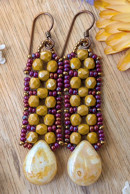 Yellow and purple beaded earrings with speckled cream teardrops at the bottom resting on a wood background.
