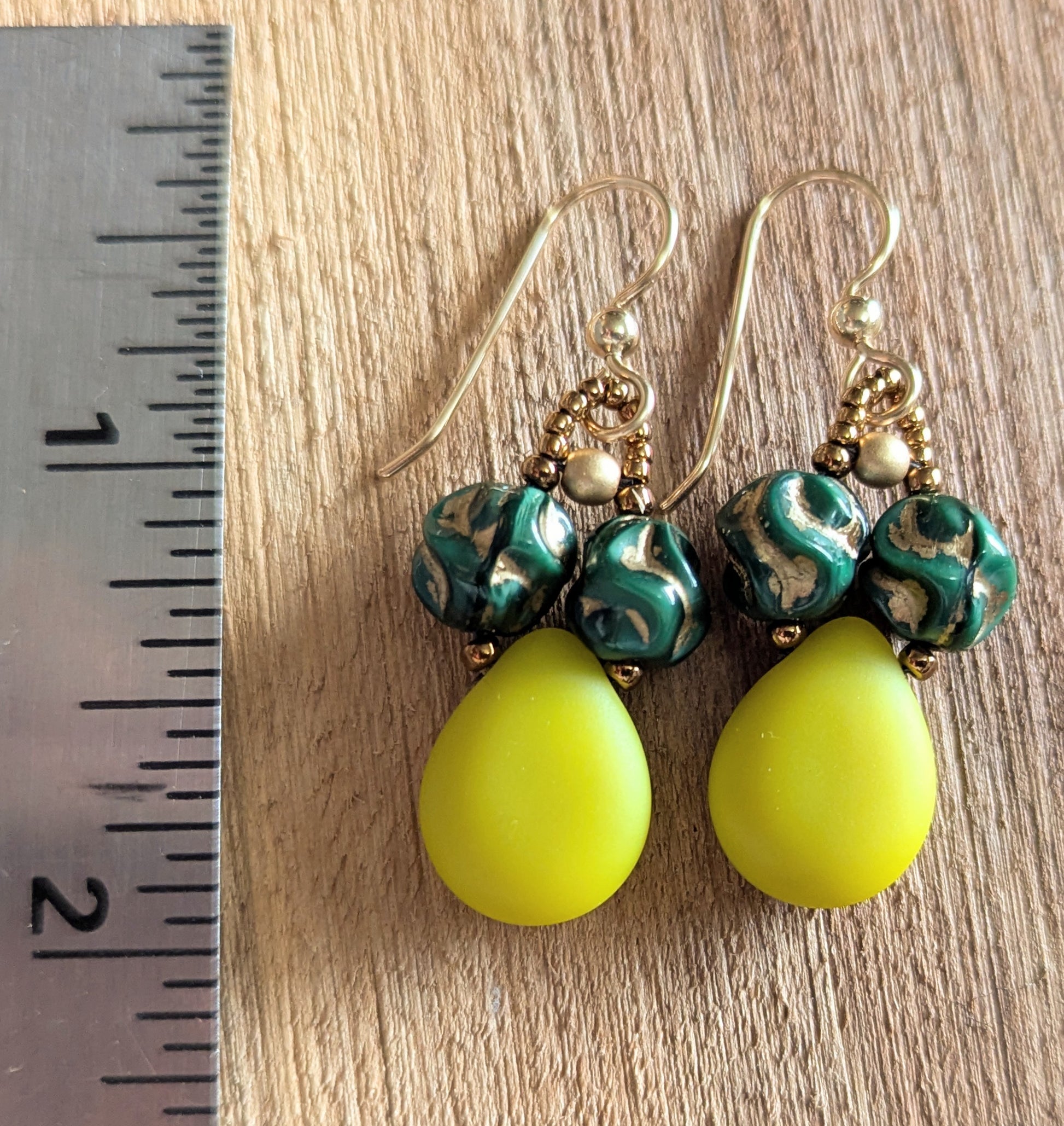 Green and yellow teardrop earrings with gold accents on a wooden surface next to a ruler.