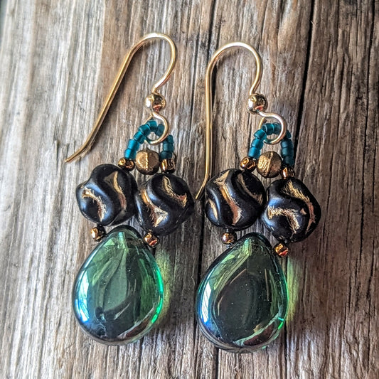 Pair of earrings with green teardrop beads and black beads with a gold pattern on a wooden surface.