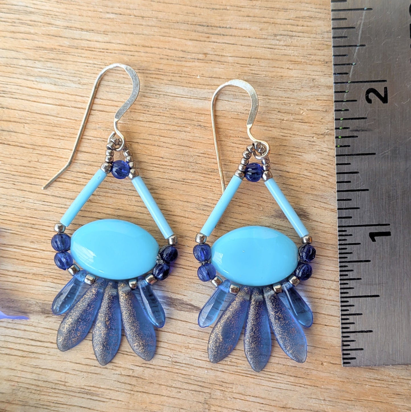 Blue and silver earrings on a wooden surface with a ruler for scale. These earrings have a light blue oval suspended by a triangle of light blue bugle beads at the top and a swag of shimmery darker blue beads around the bottom.