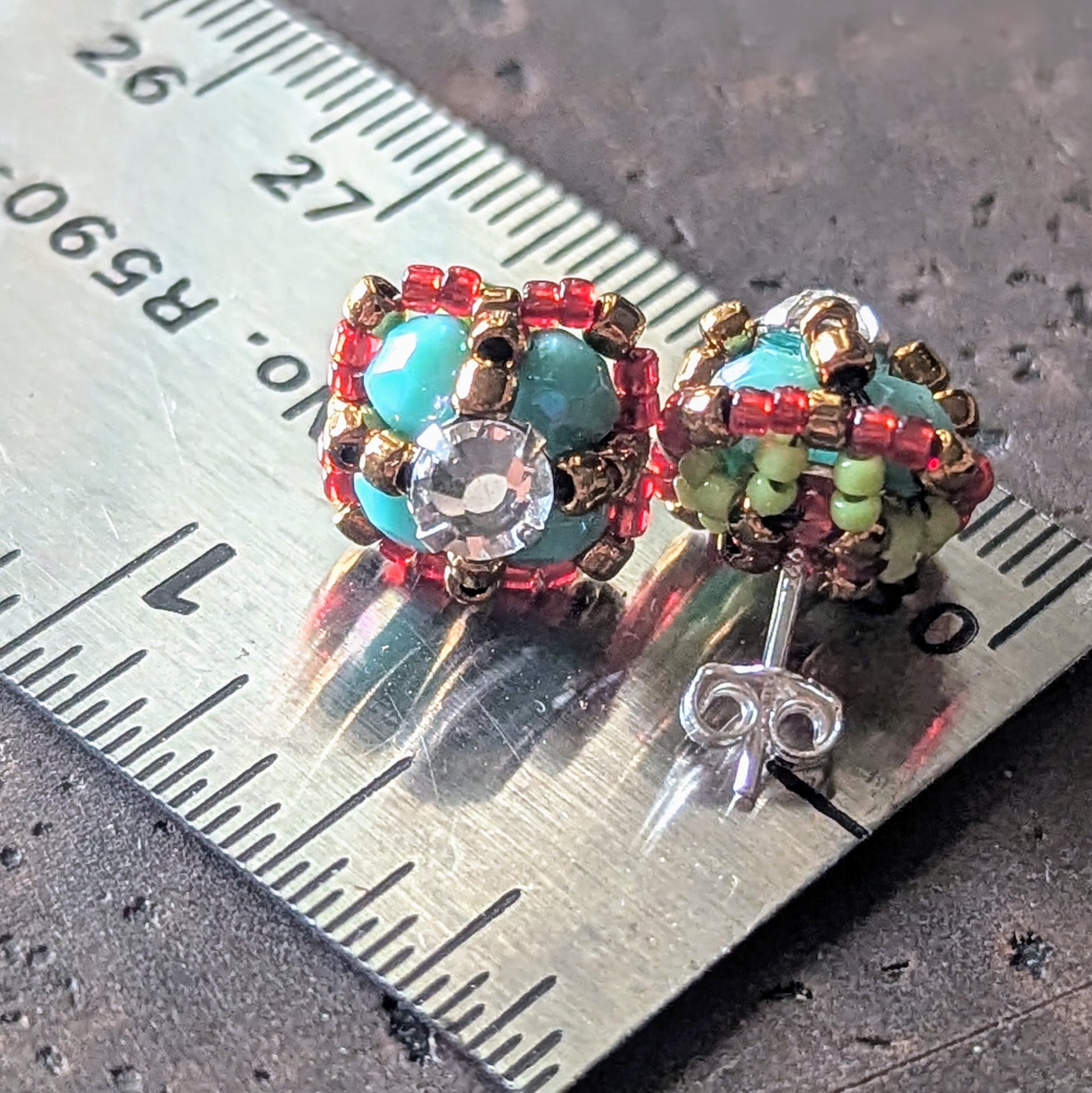 Four-lobed stud earrings on a ruler for scale. The earrings are made with larger turquoise beads surrounded by smaller red beads and a rhinestone center, shown with the back of one stud that has green details. 