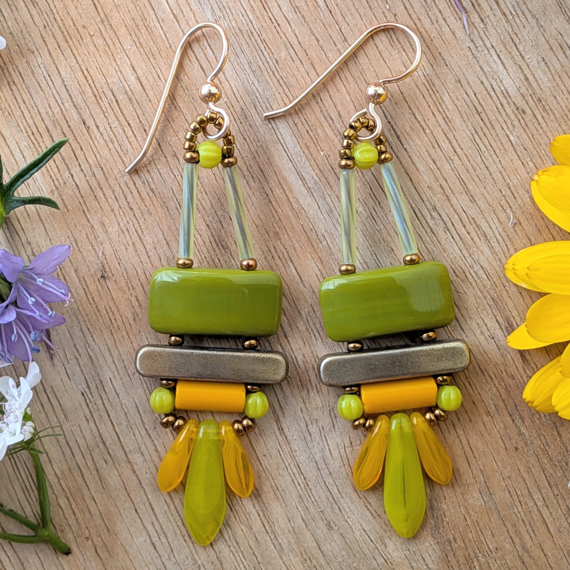 Colorful olive green, orange and chartreuse earrings on wood background. There is an avocado green rectangle on top, stacked above a gold bar. Below the bar is an orange tube and a swag of orange and chartreuse dagger beads.