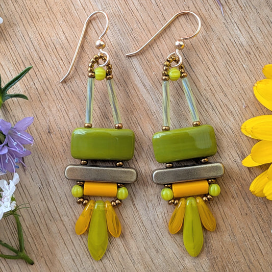 Colorful olive green, orange and chartreuse earrings on wood background. There is an avocado green rectangle on top, stacked above a gold bar. Below the bar is an orange tube and a swag of orange and chartreuse dagger beads.