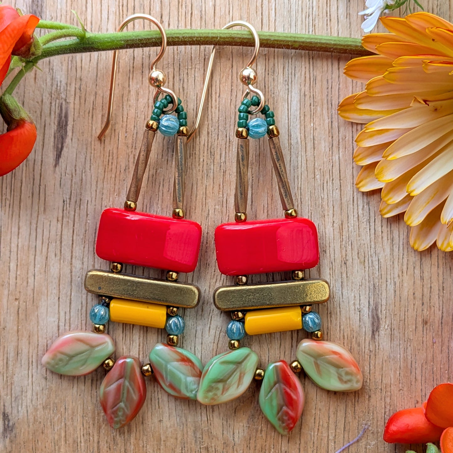 Colorful earrings on a wood background with flowers. The earrings  have a red rectangle stacked on top of a brass colored bar, with a light orange tube and swag of three red and green streaked leaves at the bottom.