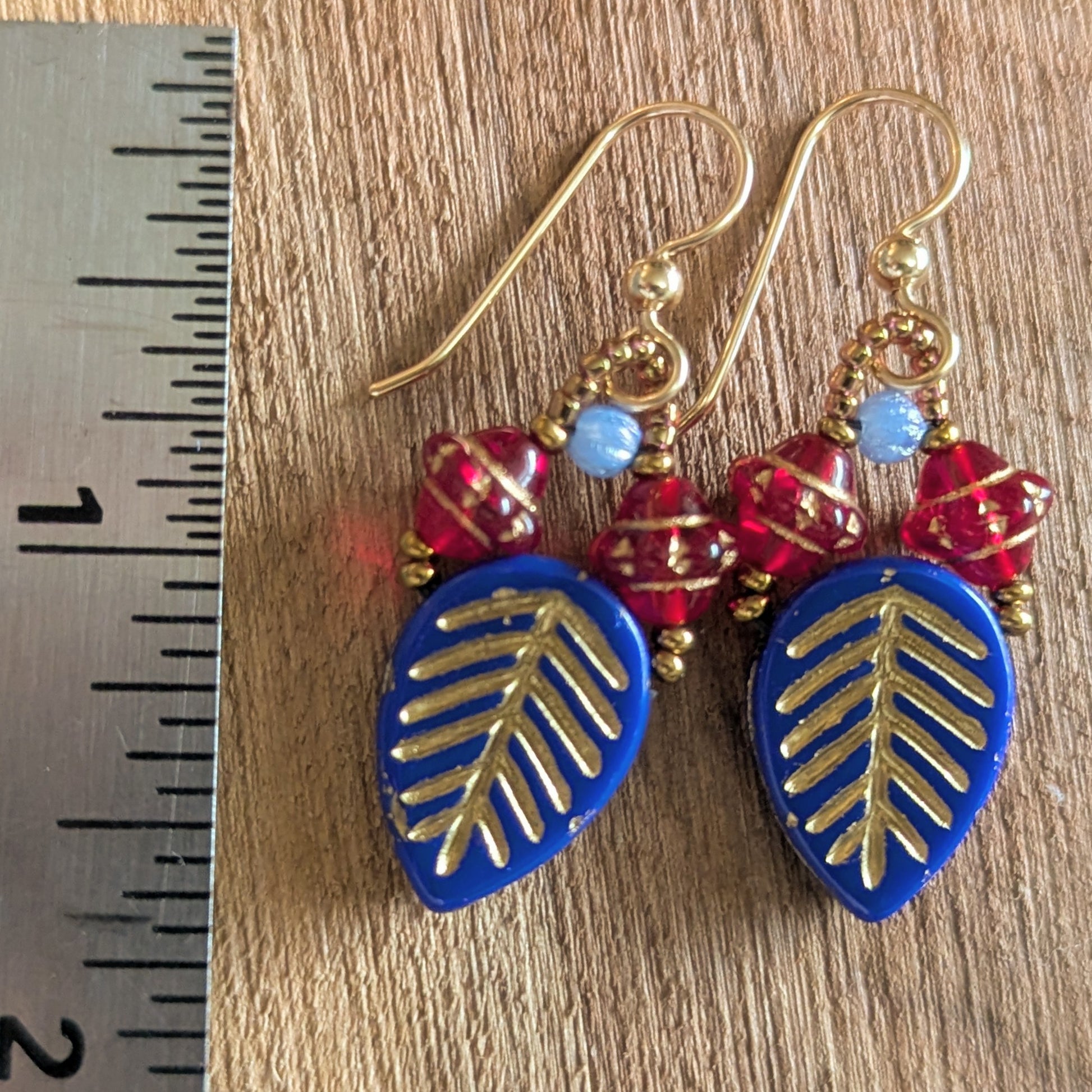 Pair of earrings with blue leaf-shaped drops, red beads, and gold accents on a wooden surface with a ruler for scale.
