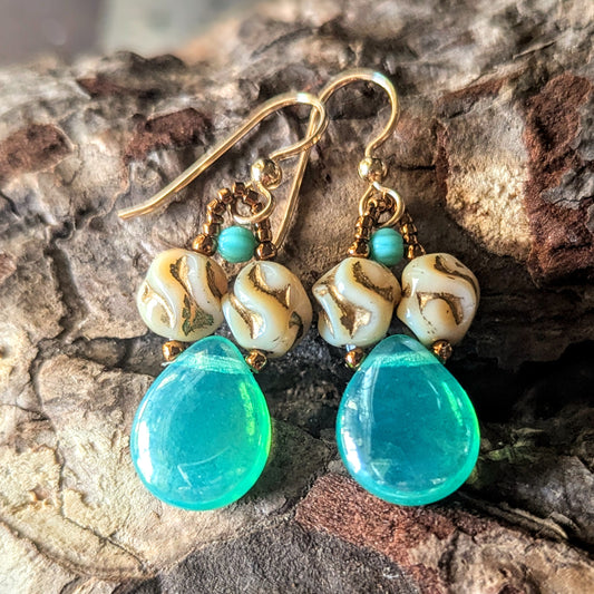 Pair of earrings with lustered blue-green teardrops and gold accents on a textured surface