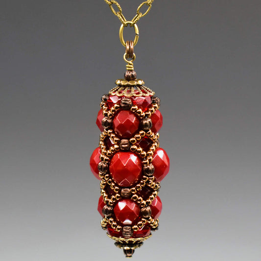 A pendant with slightly different shades of faceted red beads and gold seed bead netting overlaid hanging from a gold chain against a gray background. 