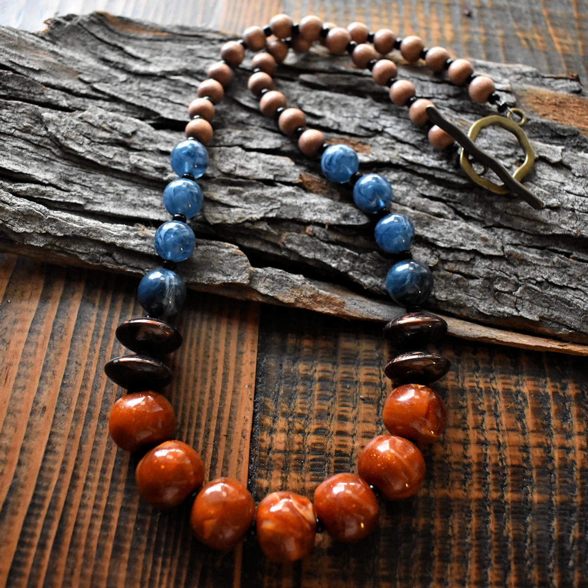 A blue and brown necklace with a large brass toggle clasp is draped across a wood background. The necklace has seven large rust colored beads with some swirls of glitter. A pair of swirled chocolate discs are on either side, followed by a series of four swirled, transparent denim blue beads. The back half of the necklace is medium brown wood beads. 
