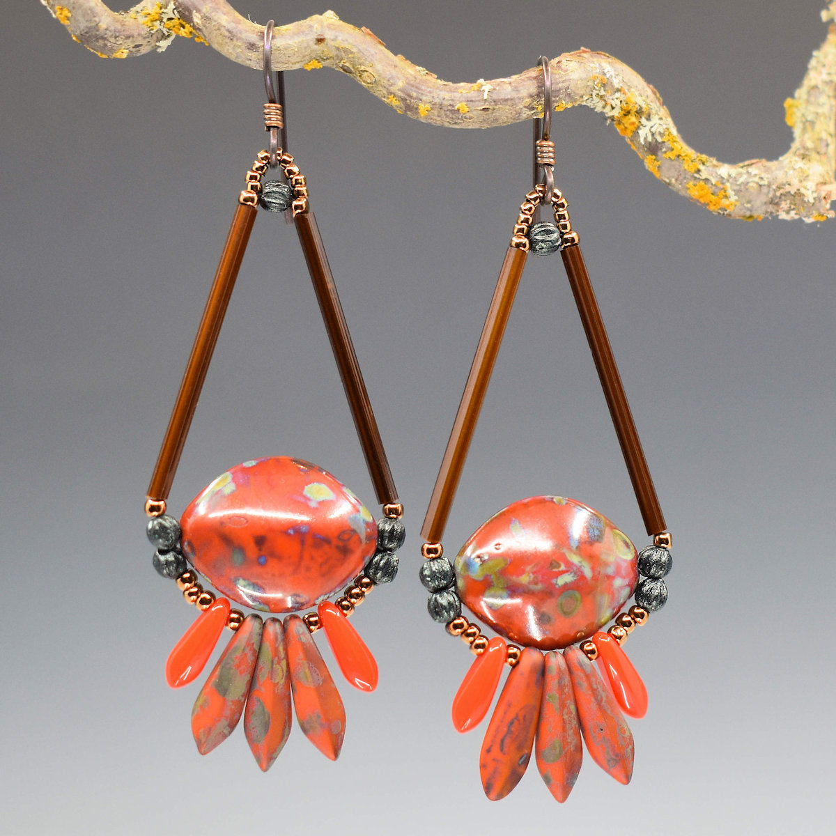 Warm red earrings hang from a lichen covered twig. The earrings are shaped like a teardrop, with long brown tube beads forming the upper sides and a warm red pointy ended oval bead with brown speckles forms the bottom. Below the teardrop shape there is a swag of red dagger beads; the inner three are larger with brown speckles and the outer are smaller and glossy. 
