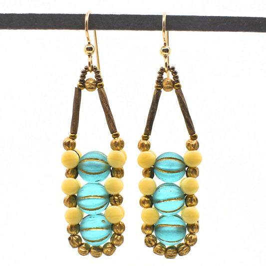 Long sea glass blue earrings with cream and gold accents white background. There are three light sea blue beads stacked vertically in the center of these earrings, like peas, surrounded by an outline of cream and gold beads. The ear wires and small accents are gold.