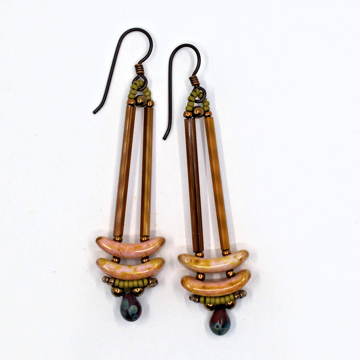 Earrings that look similar to a medicine dropper lay against a white background. There are two long, brown beads forming narrow vertical columns ending at the top of two speckled pink stacked arc shaped beads. At the bottom is a speckled red drop shaped bead, surrounded by a belt of gold seed beads.