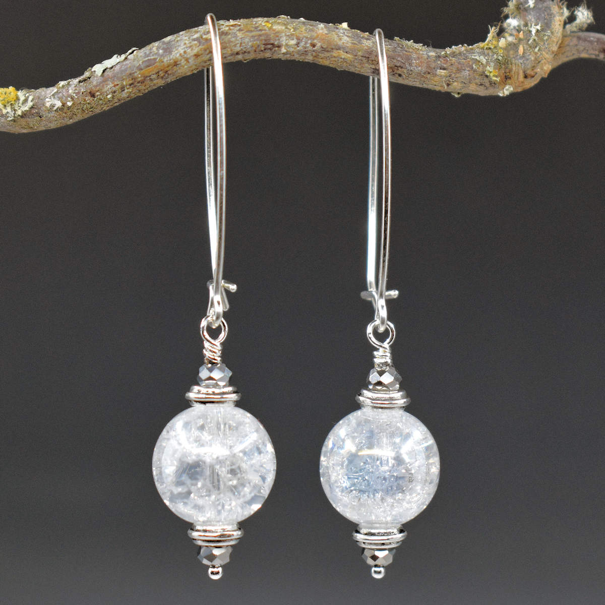 A pair of earrings hang from a twisty branch against a gray background. The earrings have long silver oval wires that latch and dangles made from shiny crackled glass balls with sparkly silver accents on the top and bottom.