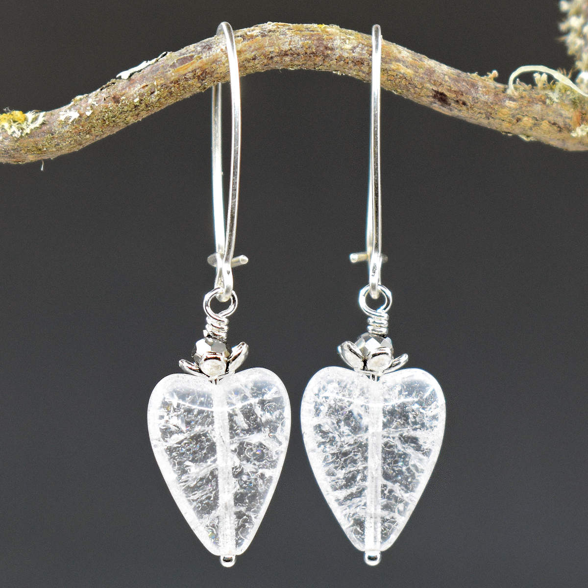 A pair of earrings hang from a twisty branch against a gray background. The earrings have long silver oval wires that latch and dangles made from shiny crackled glass hearts with sparkly silver accents on the top.