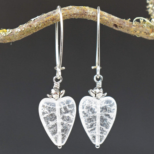 A pair of earrings hang from a twisty branch against a gray background. The earrings have long silver oval wires that latch and dangles made from shiny crackled glass hearts with sparkly silver accents on the top.