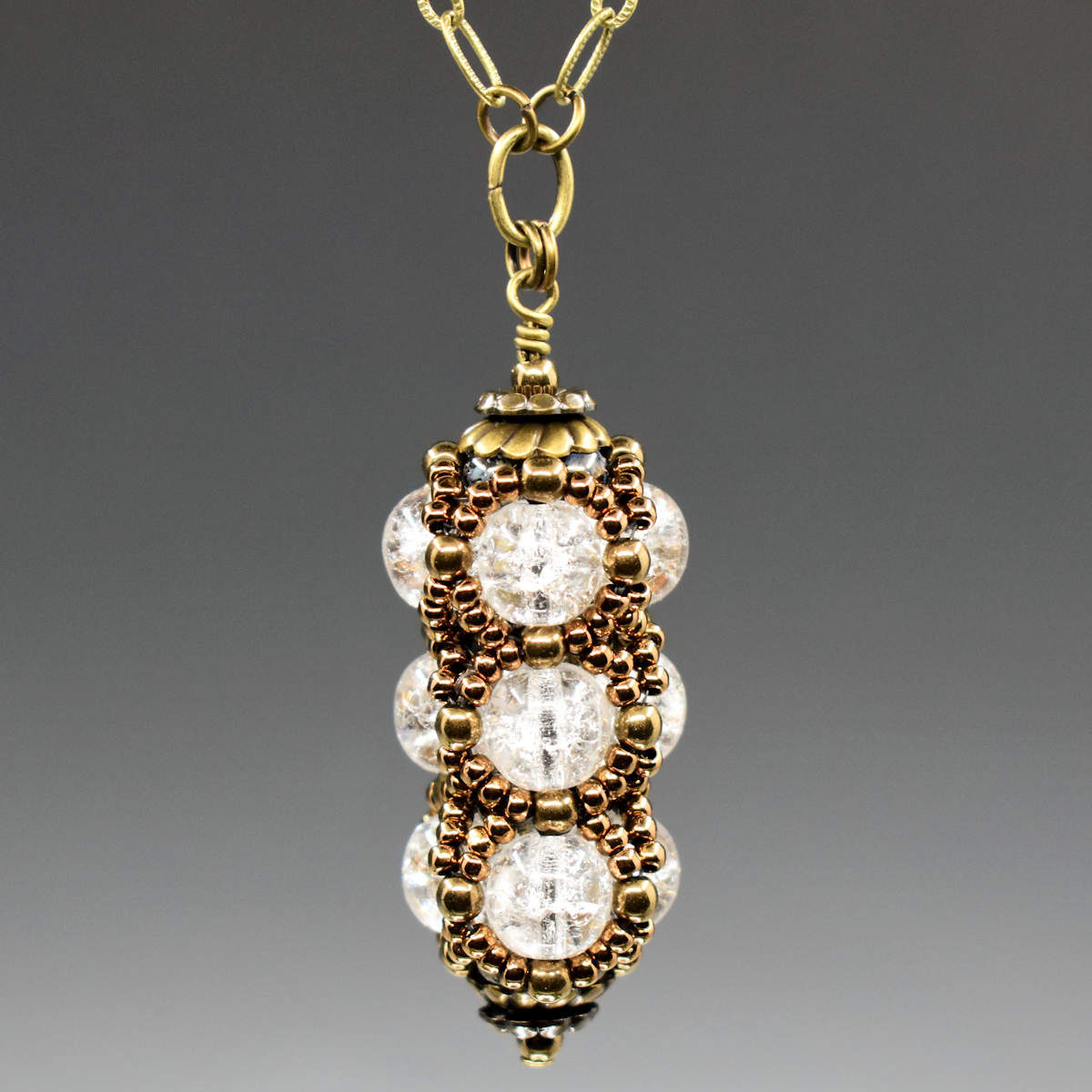 A columnar pendant formed from clear crackle glass and a netting of gold seed beads is suspended from a gold chain. The pendant has a stack of gold tone findings that cap it at the top and bottom.