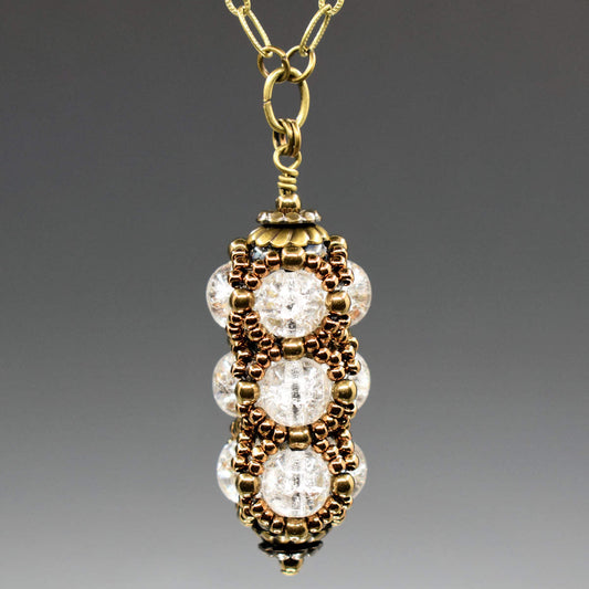 A columnar pendant formed from clear crackle glass and a netting of gold seed beads is suspended from a gold chain. The pendant has a stack of gold tone findings that cap it at the top and bottom.