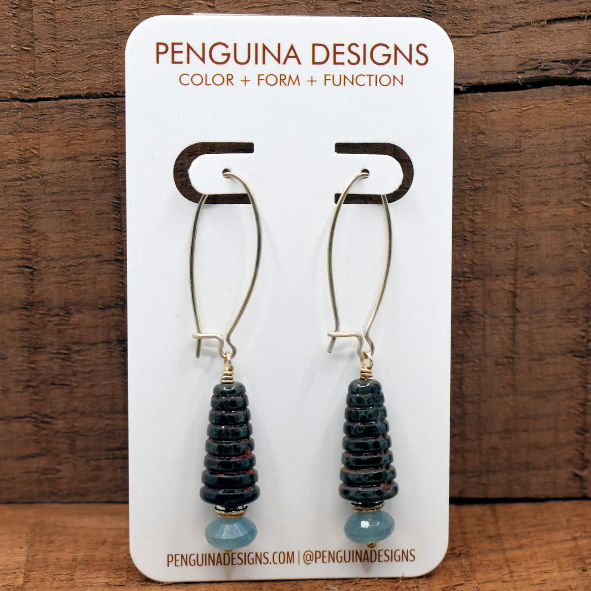 A pair of earrings on a white card rest against a wood background. The earrings have long silver oval wires that latch and  drops that are made from ridged speckled black cone beads that have smaller gray-blue rondelle beads below them.