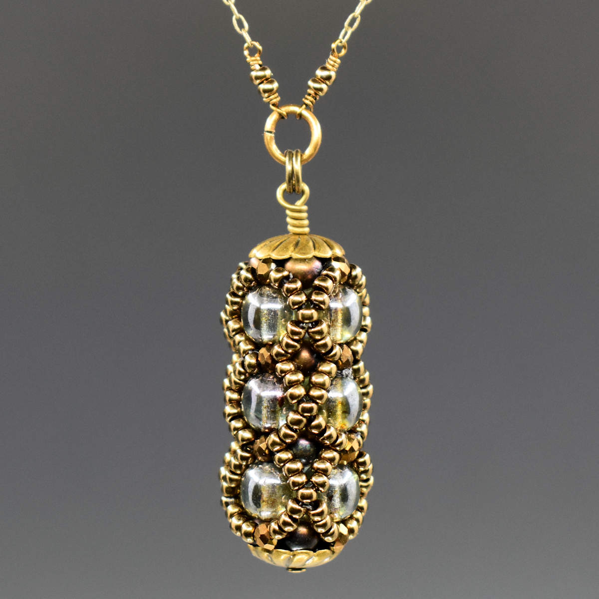 A small columnar pendant in gold and olive green hangs from a golden chain. The primary beads used are three stacked rows of lustered olive green rounds, surrounded by a netting of gold seed beads. 