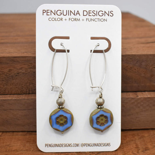 A pair of earrings on a white card rest against a wood background. The earrings have long silver oval wires that latch and drops that are made from coin-shaped periwinkle blue beads that have embossed flower medallions in their middles.
