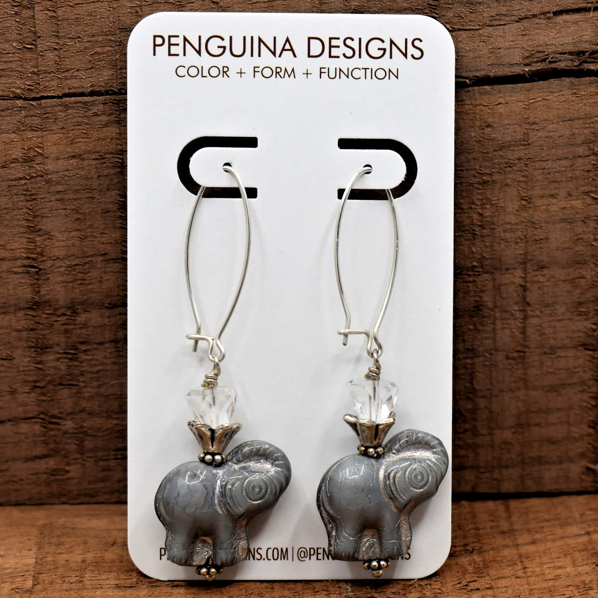 A pair of earrings on a white card rest against a wood background. The earrings have long silver oval wires that latch and drops made from chunky silvery gray elephants. Each elephant has an upturned silver cone holding a clear crystal bead on its back.