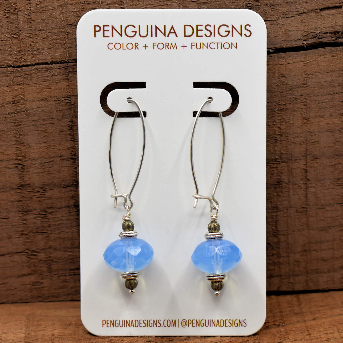 A pair of earrings on a white card rest against a wood background. The earrings have long silver oval wires that latch and  drops made from big translucent pale blue rondelle beads.