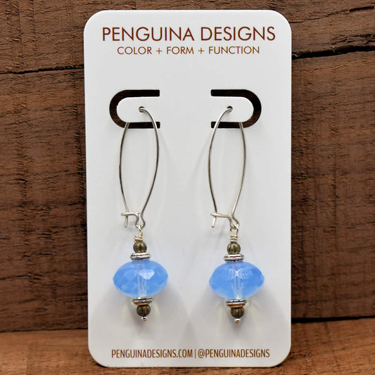 A pair of earrings on a white card rest against a wood background. The earrings have long silver oval wires that latch and  drops made from big translucent pale blue rondelle beads.