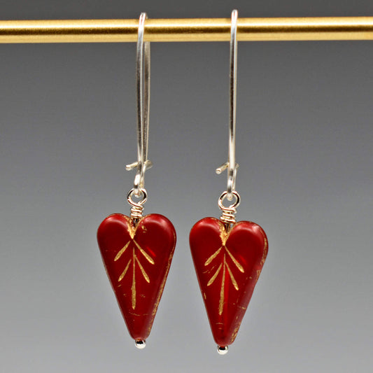 A pair of earrings on a white card hang from a gold bar, against a gray background. The earrings have long silver oval wires that latch and slender translucent red hearts at the bottom.