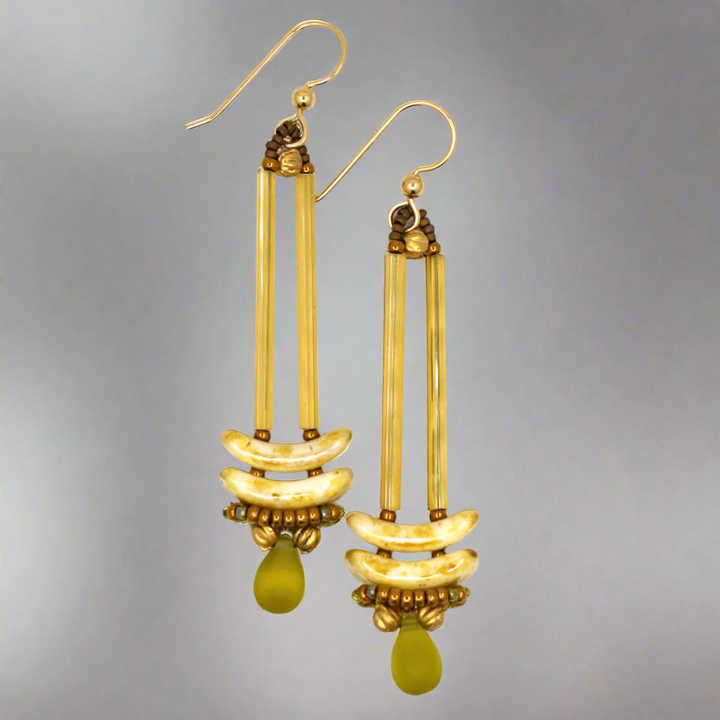 Earrings that look similar to a medicine dropper lay on a white background. There are two long, yellow-gold beads forming narrow vertical columns ending at the top of two speckled cream stacked arc shaped beads. At the bottom is a matte transparent olive green drop shaped bead, surrounded by a belt of gold seed beads.