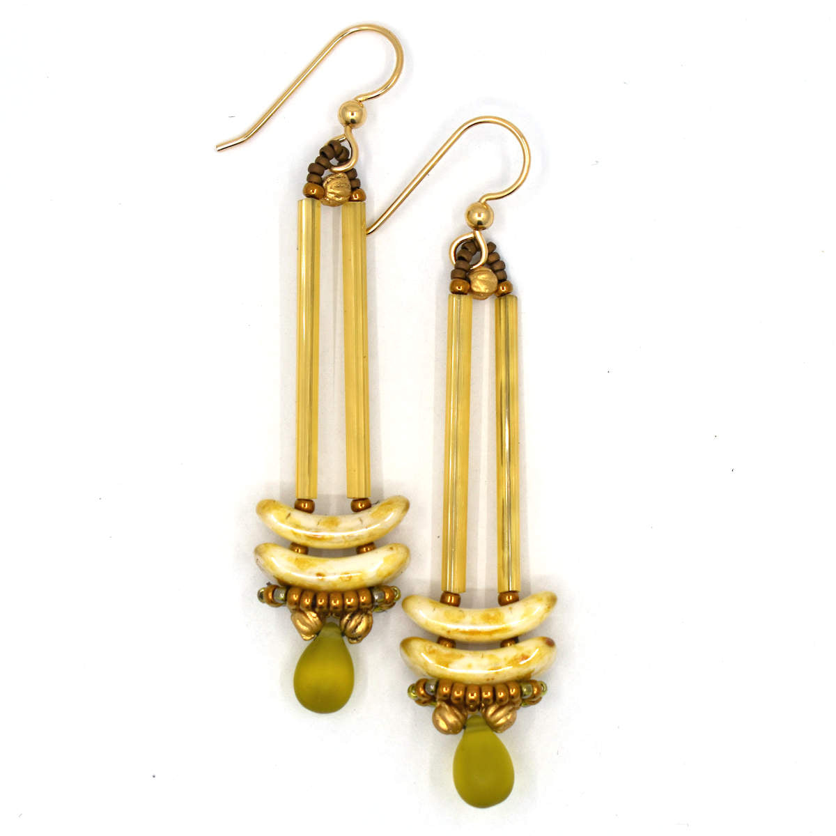 Earrings that look similar to a medicine dropper lay on a white background. There are two long, yellow-gold beads forming narrow vertical columns ending at the top of two speckled cream stacked arc shaped beads. At the bottom is a matte transparent olive green drop shaped bead, surrounded by a belt of gold seed beads.