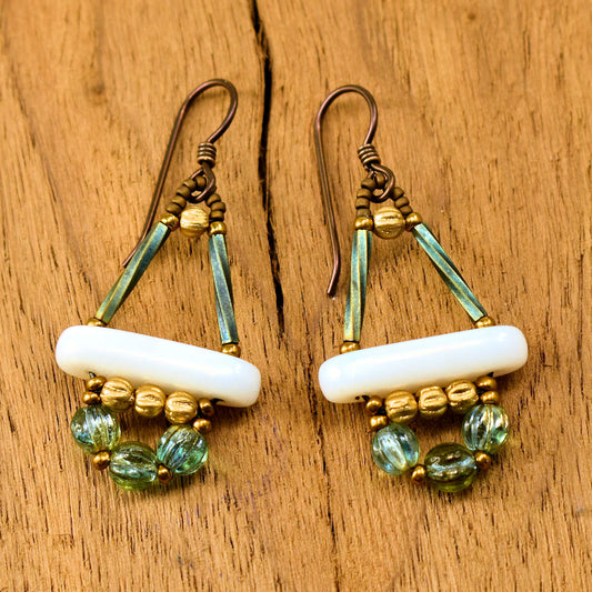 White, gold, and aqua earrings with dark ear wires lay on a wood background. These earrings are shaped like a triangle with a rocker bottom. The top of the triangle is iridescent twisted tube beads and the bottom is a white bar. Below the bar is a row of ridged round gold beads. Below that is an arc of slightly larger ridged round beads in a lustered transparent light aqua.