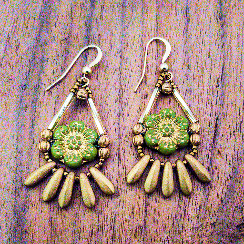 Gold earrings with a green flower in the middle lay on a wood background. The earrings have gold ear wires and are made from small gold tube and seed beads formed in a teardrop outline around avocado green flowers embossed with gold details. At the bottom of the teardrop shape there is a fringe of five small dagger beads. 
