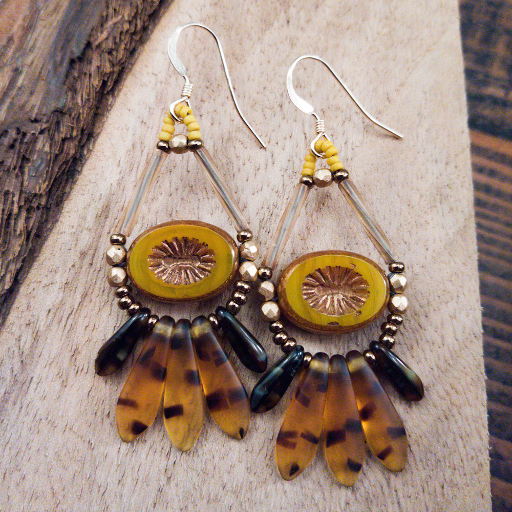 Mustard yellow and brown earrings with gold wires lay on a wood background. The earrings are formed at the top as a teardrop shape, with transparent tan tube beads and a mustard yellow oval with a starburst embossed in it's center. Below the ovals is a swag of three amber dagger beads with a tortoiseshell pattern sandwiched between two smaller black-brown variegated daggers. 