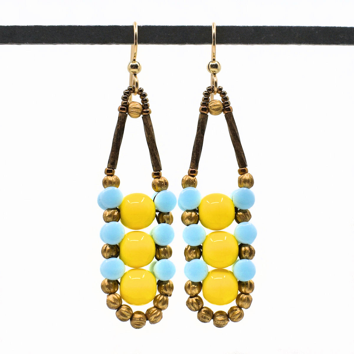 Long bright yellow and light blue earrings with gold accents and ear wires hang from a black bar in front of a white background. These earrings have three bright yellow beads stacked vertically and outlined in an alternating pattern of light blue and gold beads. 