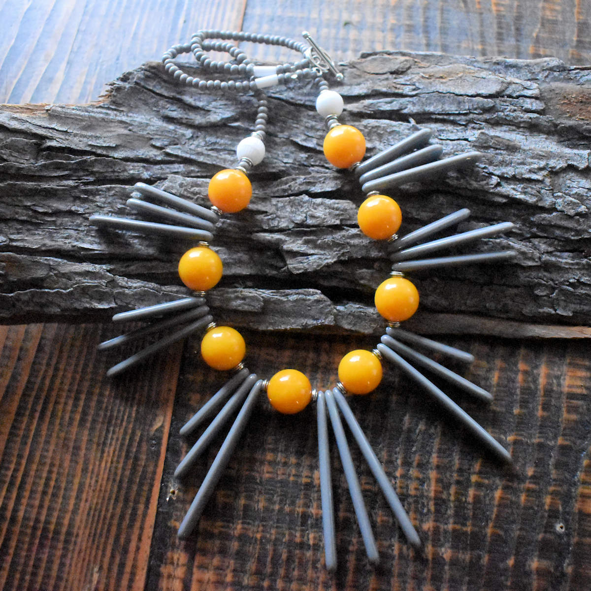 A necklace with seven clusters of long gray spines alternating with warm yellow beads is draped across a wood background. The back part of the necklace has chunky gray seed beads and a silver toggle clasp.